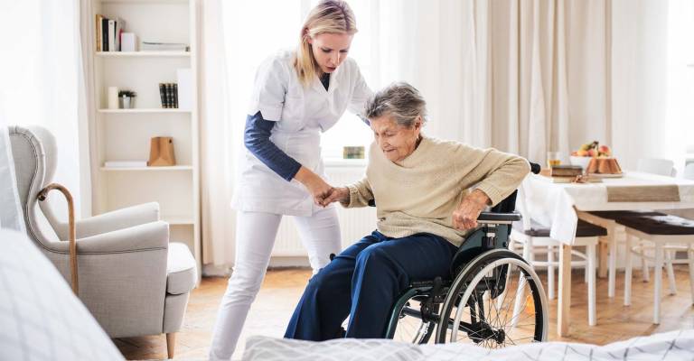 Een zorgmedewerkster helpt een oude dame op te staan vanuit haar rolstoel